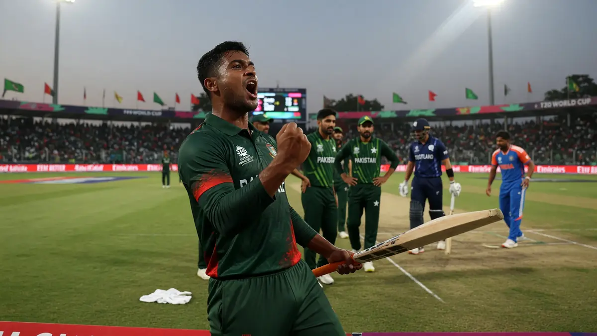 Cricketer in a dark green kit celebrating with a raised fist and bat under stadium lights, with teammates and opponents standing on the field behind him